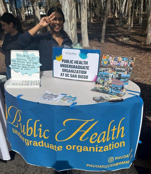 A table at Triton Day 2026 with signage for the Public Health Undergraduate Organization, photos from org events, and chapstick. A handwritten sign near the chapstick reads: Health lips help protect your body! Cracked lips make it easier for bacteria and viruses to enter!" Behind the table stands a female college student, one of the PHUO presidents, holding up 2 fingers in the "peace" sign.